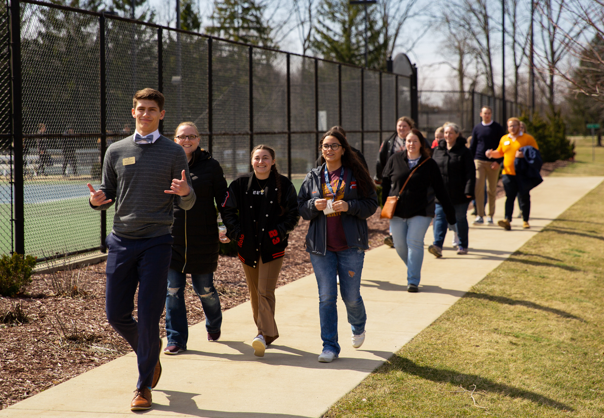 An admissions intern giving a campus tour walking past the tennis cours with students smiling.