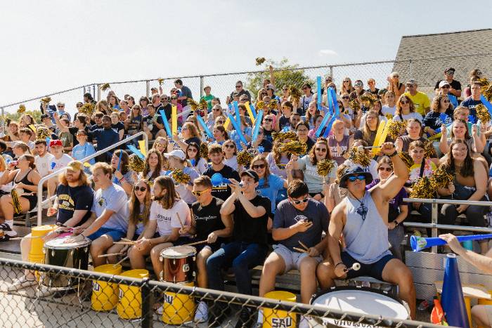 Students cheering in the stands at the soccer game on a bright, sunny day.