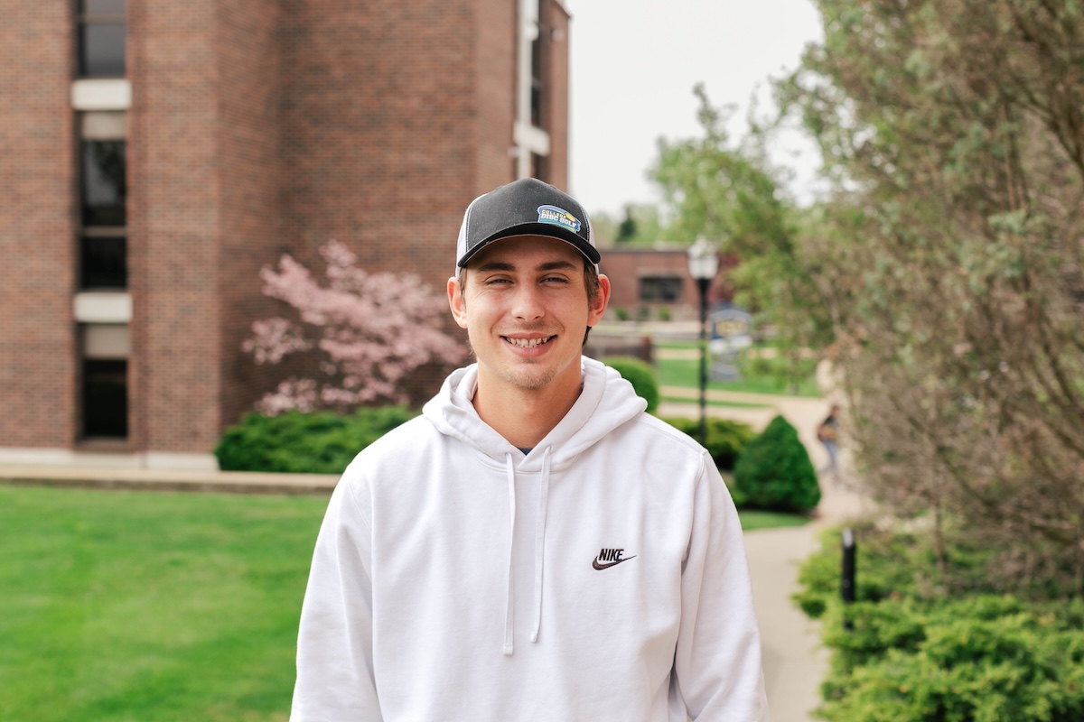 A man wearing a baseball cap standing facing the camera with a building and some greenery in the background