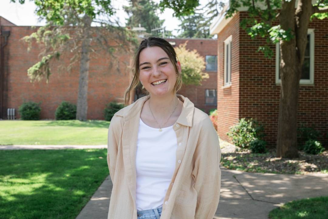 A female standing outside smiling at the camera. There is a red brick building in the background as weel as grass and trees